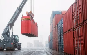 Wózek widłowy z wysięgnikiem podnosi czerwony kontener w mglistym porcie. Po prawej stos kontenerów.