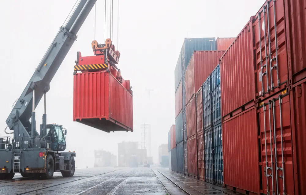 Wózek widłowy z wysięgnikiem podnosi czerwony kontener w mglistym porcie. Po prawej stos kontenerów.